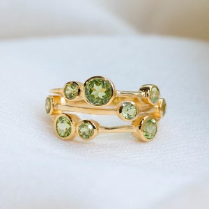 May include: Two gold rings with multiple round, light green gemstones. The rings are stacked on top of each other, with the gemstones varying in size. The rings are set against a white, textured background.