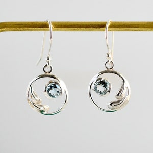 May include: Silver earrings with a delicate leaf design and a light blue gemstone in the center of each circle.