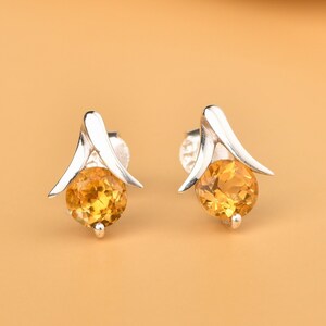 May include: A pair of silver earrings with a small, round, yellow gemstone in the center of each earring. The earrings have a delicate, leaf-like design.