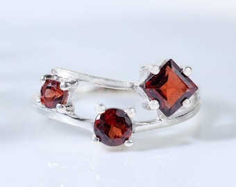 May include: A silver ring with three red gemstones. The ring has a unique design with two bands that intertwine and feature the gemstones.