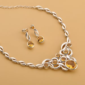 Op de afbeelding: Een zilveren ketting- en oorbellenset met ovale, amberkleurige edelstenen. De ketting heeft een decoratief ontwerp met meerdere edelstenen en een ketting. De oorbellen hebben elk een enkele edelsteen. De sieradenset staat op een beige achtergrond.