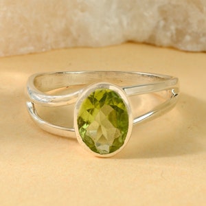 Op de afbeelding: Een zilveren ring met een split-schacht ontwerp, met een ovaal geslepen, lichtgroene edelsteen. De edelsteen is het middelpunt, gezet in een bezel setting. De ring heeft een moderne, minimalistische stijl.