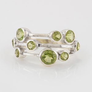 Può includere: Un anello d'argento con più bande e pietre preziose verdi. L'anello ha un design moderno con un gruppo di pietre rotonde e ovali.