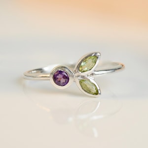 Può includere: Un anello d'argento con una pietra preziosa di ametista viola e due pietre preziose di peridoto verde a forma di foglie.