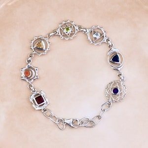 Puede incluir: Pulsera de plata con siete dijes, cada uno representando un chakra con una piedra preciosa de color diferente. Los dijes están conectados por una cadena de plata.