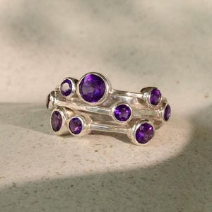 May include: A silver ring featuring multiple round, deep purple gemstones. The ring has a unique design with the gemstones set in a cluster arrangement. The ring is displayed on a light-colored, textured surface, showcasing its intricate details and elegant style.