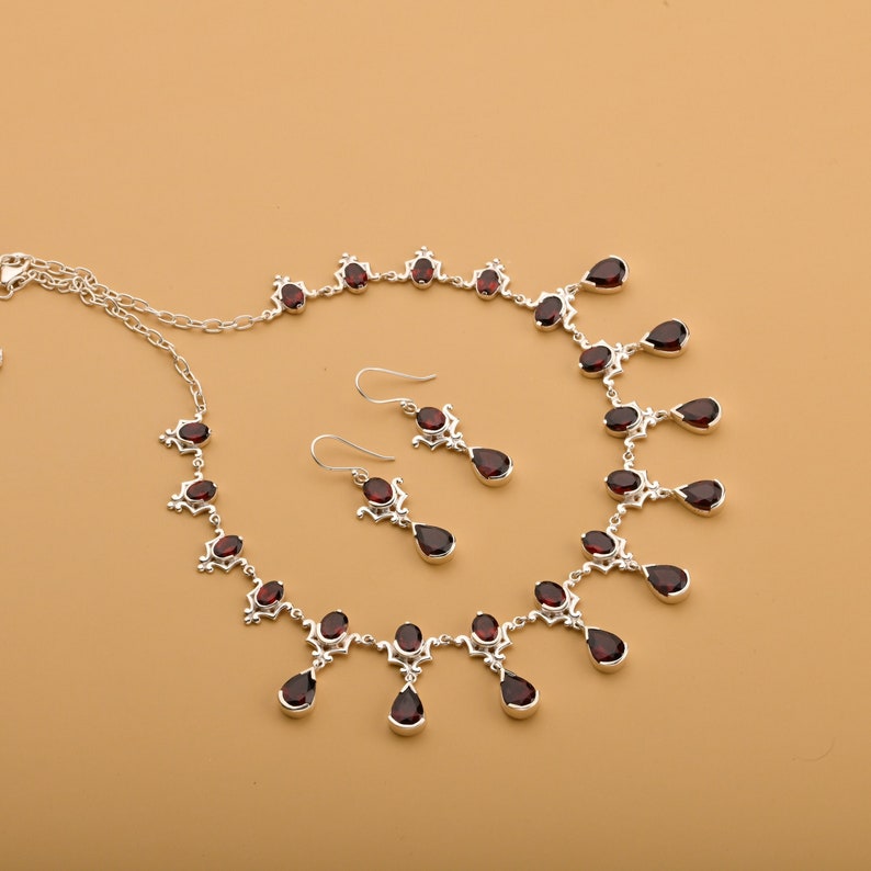 Puede incluir: Un collar y pendientes de plata con gotas de piedras preciosas rojas. El collar tiene una cadena delicada con un dise&ntilde;o de colgante con m&uacute;ltiples gotas de piedras preciosas rojas.