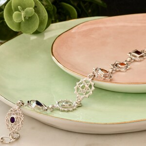 May include: A silver bracelet with seven chakra symbols and gemstones. Each symbol is a circle with a star inside and a gemstone in the center. The gemstones are amethyst, lapis lazuli, turquoise, green aventurine, citrine, carnelian, and red jasper.