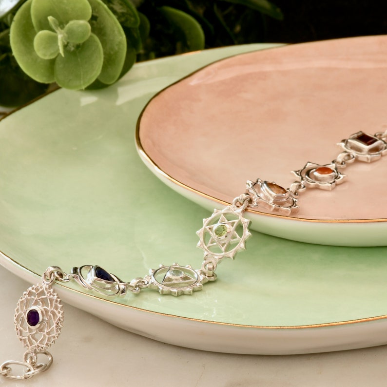 May include: A silver bracelet with seven chakra symbols and gemstones. Each symbol is a circle with a star inside and a gemstone in the center. The gemstones are amethyst, lapis lazuli, turquoise, green aventurine, citrine, carnelian, and red jasper.
