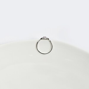 Op de afbeelding: Een zilveren ring met een kleine roze steen in het midden. De ring ligt op een wit oppervlak.
