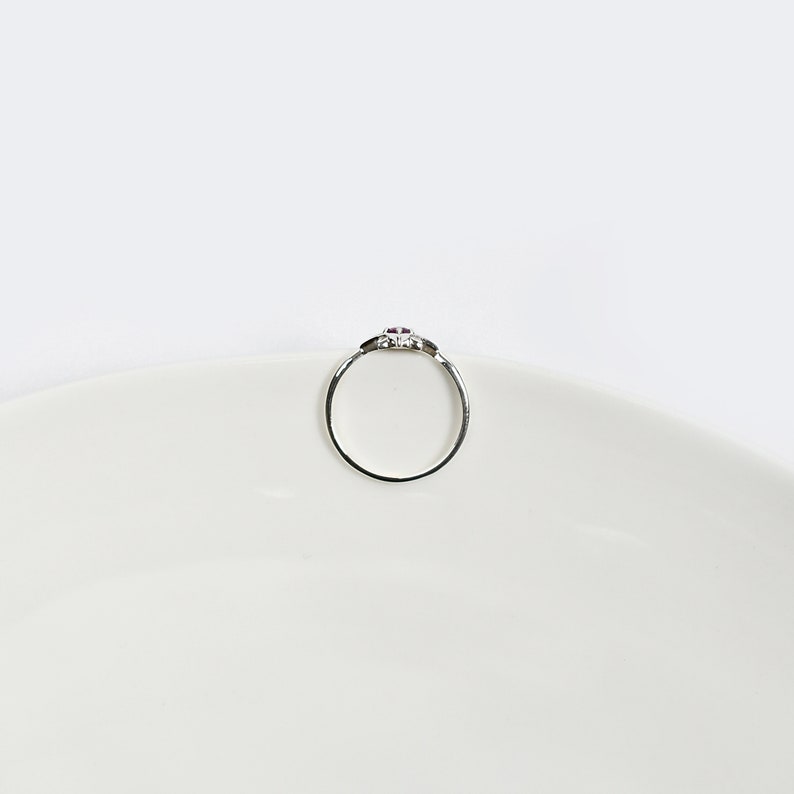 Op de afbeelding: Een zilveren ring met een kleine roze steen in het midden. De ring ligt op een wit oppervlak.