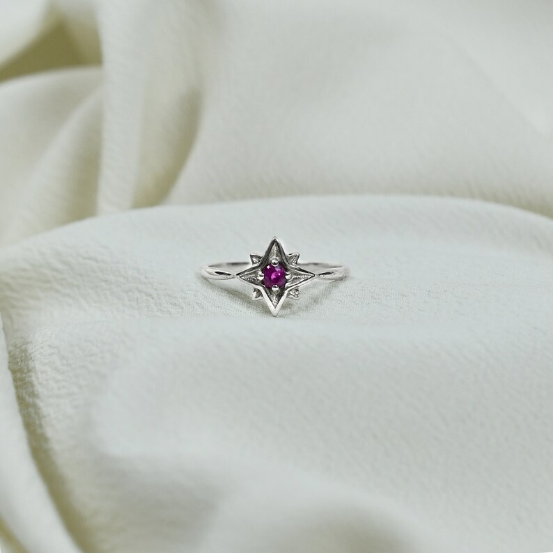 Op de afbeelding: Een zilveren ring met een stervormig ontwerp met een kleine, ronde, roze edelsteen in het midden.