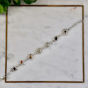 May include: Silver bracelet with seven chakra symbols and gemstones. Each symbol is a silver mandala with a gemstone in the center. The gemstones are red, orange, yellow, green, blue, indigo, and violet.