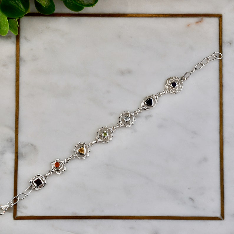 May include: Silver bracelet with seven chakra symbols and gemstones. Each symbol is a silver mandala with a gemstone in the center. The gemstones are red, orange, yellow, green, blue, indigo, and violet.