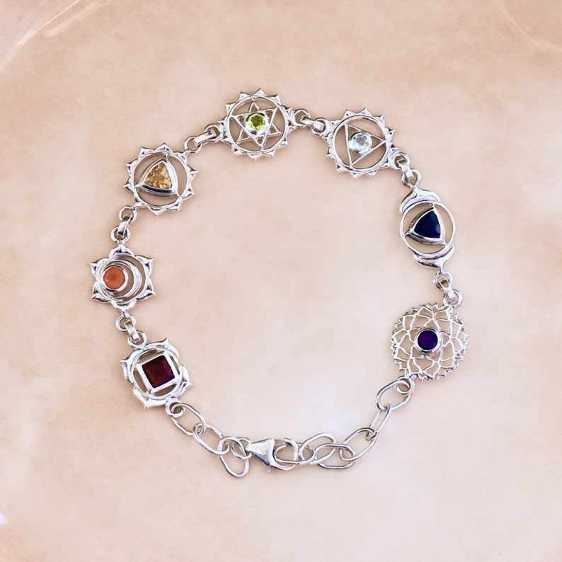 May include: Silver bracelet with seven charms, each representing a chakra with a different colored gemstone. The charms are connected by a silver chain.