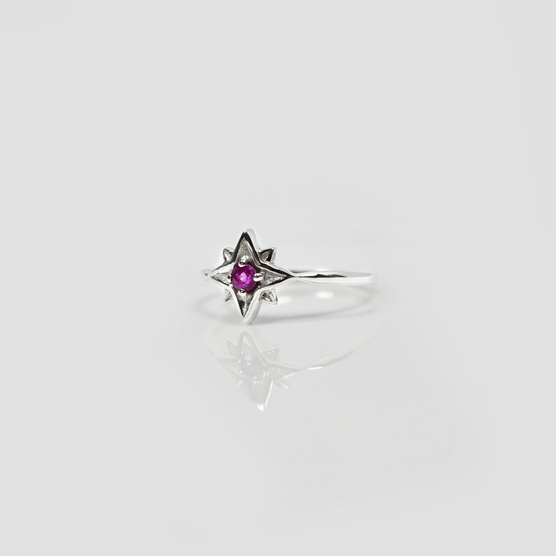 Op de afbeelding: Een zilveren ring met een roze edelsteen in het midden van een stervormig ontwerp.