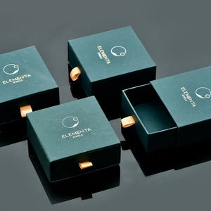 May include: Four green boxes with a gold logo that says "Elementa Jewels" and a gold pull tab. The boxes are on a black surface.