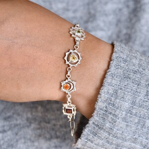 May include: A silver bracelet with seven chakra symbols and gemstones. The symbols are in a circular shape and the gemstones are in the center of each symbol. The bracelet is on a person's wrist.