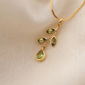 May include: A gold necklace with a pendant shaped like a branch with green gemstone leaves. The pendant has a teardrop-shaped gemstone at the bottom.