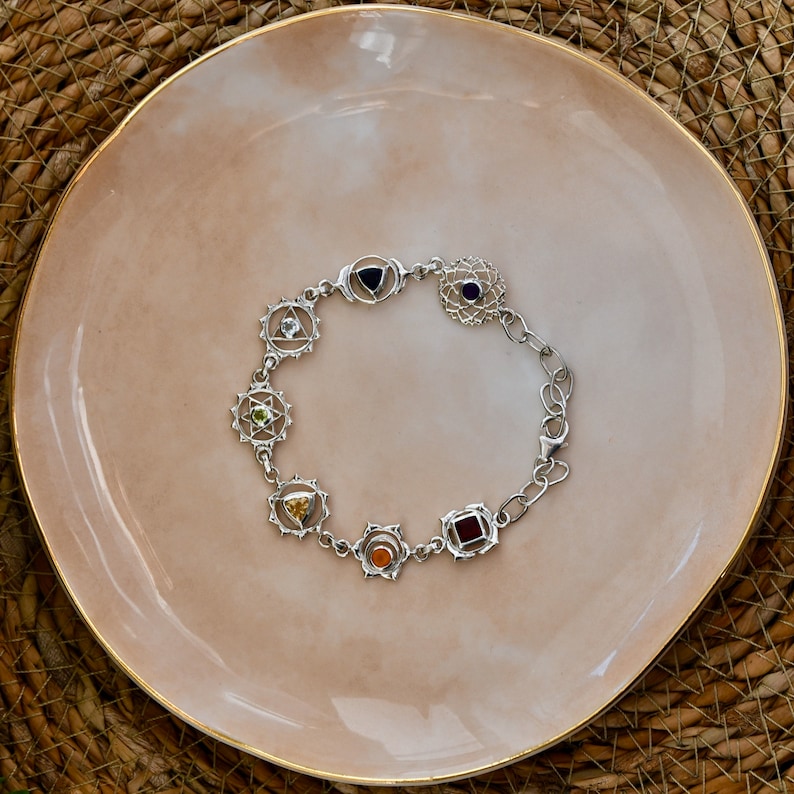 May include: A silver bracelet with seven charms, each featuring a different colored gemstone and a geometric design. The bracelet is on a light pink plate.