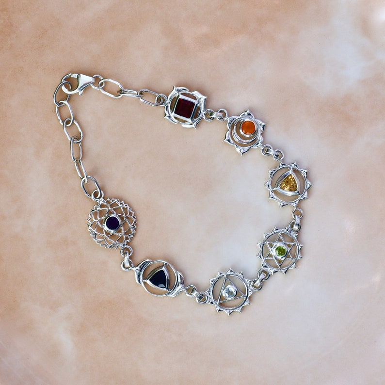 May include: Silver bracelet with seven chakra symbols, each with a different colored gemstone. The gemstones are red, orange, yellow, green, blue, indigo, and violet.