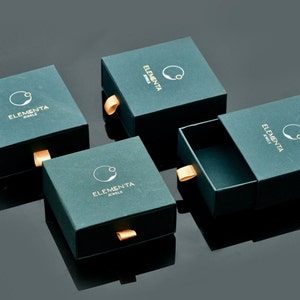 Peut inclure: Quatre bo&icirc;tes vert fonc&eacute; avec des accents dor&eacute;s et le texte "Elementa Jewels" sur chaque bo&icirc;te. Les bo&icirc;tes sont dispos&eacute;es en quinconce sur une surface noire.