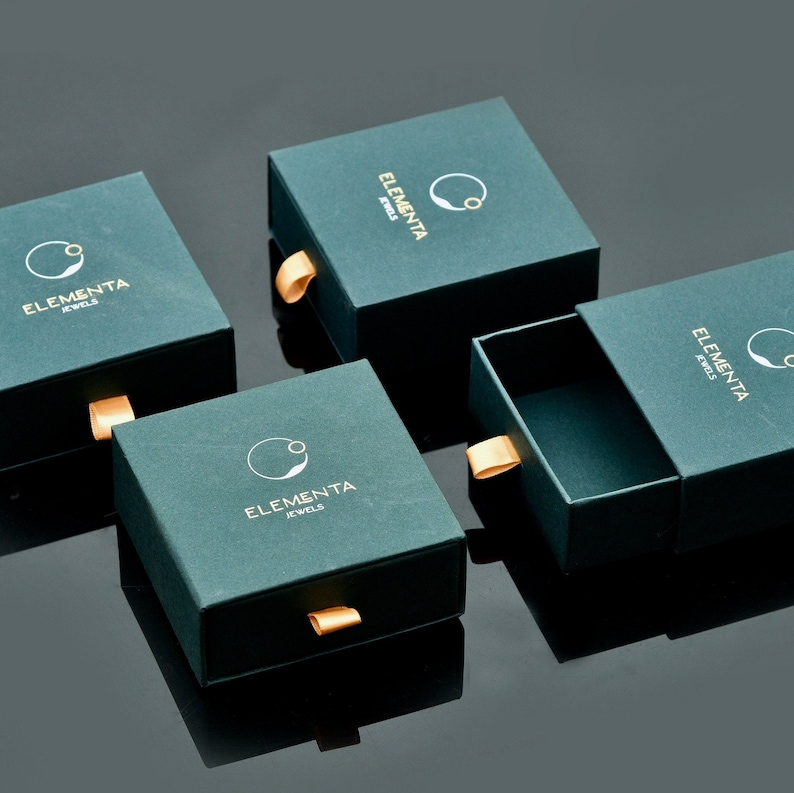 Peut inclure: Quatre bo&icirc;tes vert fonc&eacute; avec des accents dor&eacute;s et le texte "Elementa Jewels" sur chaque bo&icirc;te. Les bo&icirc;tes sont dispos&eacute;es en quinconce sur une surface noire.