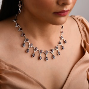 Puede incluir: Un collar de plata con un colgante de granate en forma de l&aacute;grima. El collar tiene una cadena delicada y est&aacute; adornado con varias piedras de granate m&aacute;s peque&ntilde;as.