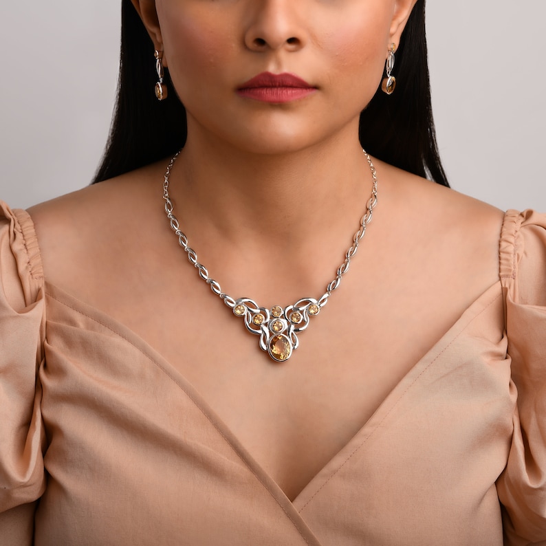 Op de afbeelding: Een zilverkleurige ketting- en oorbellenset met geel-oranje edelstenen. De ketting heeft een centrale hanger met een grote ovale steen en kleinere stenen. De oorbellen hebben een vergelijkbaar ontwerp.