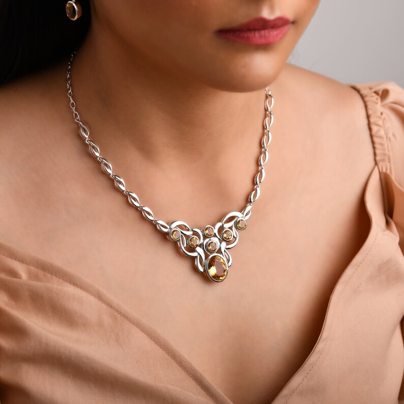 Op de afbeelding: Zilverkleurige ketting en bijpassende oorstekers met een reeks ovale schakels en een centrale hanger. De hanger en oorbellen tonen ovale en ronde, amberkleurige edelstenen. De sieraden worden gedragen.