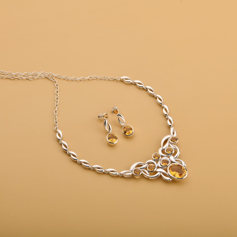 Op de afbeelding: Een zilveren ketting- en oorbellenset met ovale, amberkleurige edelstenen. De ketting heeft een decoratief ontwerp met meerdere edelstenen en een ketting. De oorbellen hebben elk een enkele edelsteen. De sieradenset staat op een beige achtergrond.