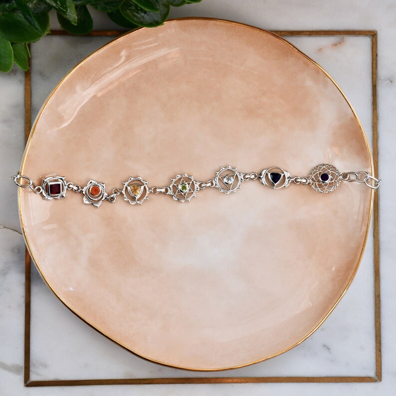May include: A silver bracelet with seven charms, each representing a chakra. The charms are decorated with colorful gemstones and intricate designs.