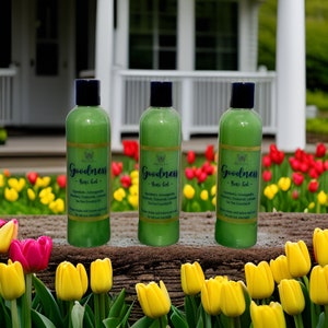 May include: Three green bottles with white labels and the text "Goodness - Yoni Gel -" on a wooden surface with yellow and pink tulips in the background.