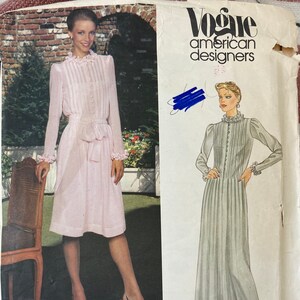 May include: Vogue American Designers sewing pattern. The pattern features a light pink dress with long sleeves, a button-down bodice, and a matching belt. The pattern also includes a drawing of a grey dress with similar features. The pattern is labeled "Albert Nipon".