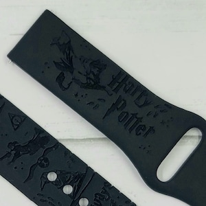 May include: Black silicone watch band with an embossed design featuring Harry Potter characters and symbols, including the Hogwarts Express, a snitch, and the Hogwarts crest. The band also includes the text "Harry Potter" and "9 3/4".
