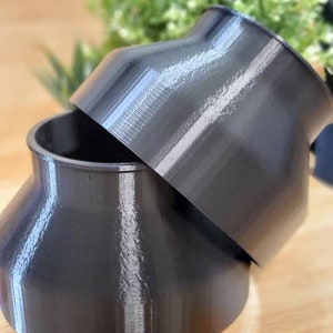 May include: Two black plastic 3D printed funnel shaped objects stacked on top of each other on a wooden surface.