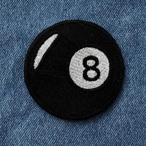 May include: A circular embroidered patch featuring a black billiard ball design. The patch has a white centre circle with a black number eight and a white curved highlight on the upper left side, all bordered by a thick black stitched edge.