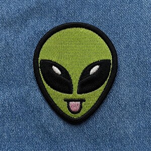 May include: An embroidered patch featuring a lime green alien head with a black border. The alien has large, almond-shaped black eyes with white highlights and a small mouth with a pink tongue sticking out, centred on a blue denim fabric.