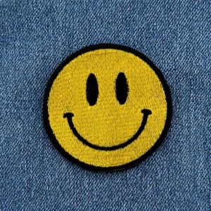 Peut inclure: Un écusson brodé circulaire présentant un motif de visage souriant jaune classique avec deux yeux ovales noirs et une large bouche incurvée noire, le tout souligné par une épaisse bordure noire sur un fond en tissu denim bleu texturé.