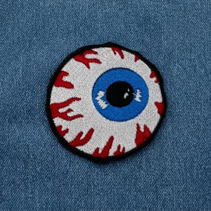 May include: A circular embroidered patch featuring a realistic eyeball design. The patch has a white sclera with red bloodshot veins, a bright blue iris, and a solid black pupil, all outlined with a thick black border on a blue denim fabric.