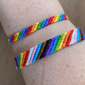May include: Two colourful woven friendship bracelets. One bracelet features a rainbow pattern, while the other includes the colours of the transgender pride flag. The bracelets are worn on a wrist.