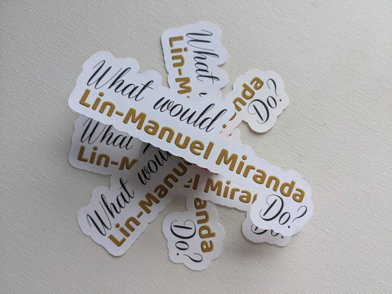 Puede incluir: Un conjunto de pegatinas blancas con letras doradas que dicen "What would Lin-Manuel Miranda Do?" Las pegatinas est&aacute;n apiladas sobre una superficie blanca.