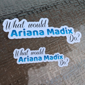 Könnte beinhalten: Zwei weiße Aufkleber mit dem Text "What would Ariana Madix Do?" in Schwarz und Blau. Die Aufkleber befinden sich auf einer strukturierten, braunen Oberfläche. Der Text ist in einer Schreibschrift.