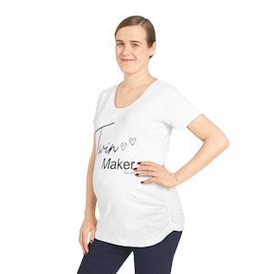 May include: White maternity t-shirt with a scoop neck. The shirt features the text "Twin Maker" in a script font, with two small heart graphics and the text "Est. 2023" below. The shirt has a ruched side detail.