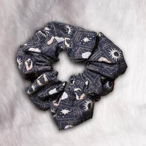 May include: A dark blue scrunchie with a celestial pattern featuring suns, stars, and constellations in white and tan. The fabric is gathered, creating a voluminous, circular shape. This hair accessory is suitable for various hairstyles.