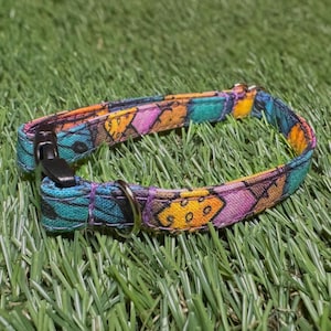 May include: A colorful pet collar with a black buckle and a silver metal ring. The collar features a patchwork design with teal, orange, yellow, and purple geometric shapes. The collar is resting on green grass.