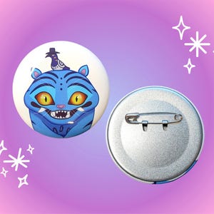 May include: Two round, blue and white pinback buttons. One button features a cartoon blue tiger with yellow eyes and a bird wearing a hat. The other button is silver with a safety pin clasp.