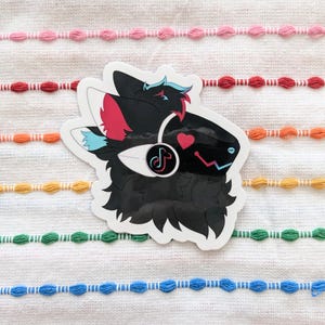 May include: A die-cut sticker featuring a stylized black creature with fluffy fur, large ears with pink and blue accents, and a dark visor. The character wears white headphones displaying a TikTok logo, a pink heart, and a blue speech bubble icon.