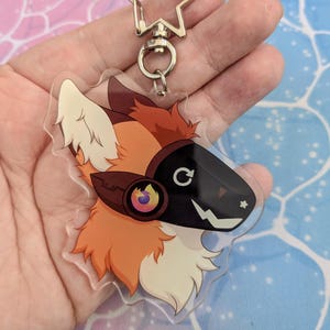 May include: A clear acrylic keychain featuring a cartoon fox with orange fur, a black mask with a white lightning bolt, and a red and yellow flame design on the headphones.