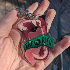 May include: A clear acrylic keychain with a cartoon image of a pink and red mouth with sharp teeth and a green tongue. The tongue is sticking out and has a green banner that says "Eat the Rich".